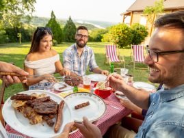 Group of friends having an outdoor barbecue lunch, eating grilled meat, drinking beer and having fun. Focus on the meat